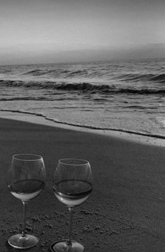 two wine glasses sitting on top of a sandy beach next to the ocean with waves coming in