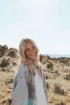 a woman standing in the middle of a field wearing a jean jacket and smiling at the camera