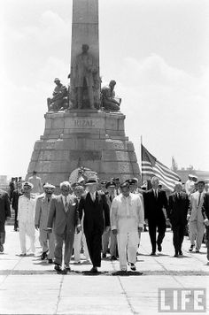 black and white photograph of men standing in front of a statue with flags on it