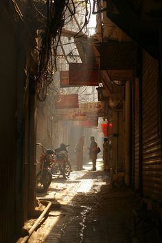 people walking down an alley way with motorcycles parked on the side walk and signs hanging above them