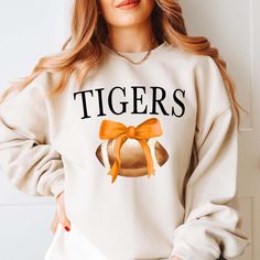 Hey there, football fan! Ready to cheer on your favorite team in style? Our "Custom Football Team with Bow" Graphic Sweatshirt is the perfect way to show off your team spirit while keeping things cute and cozy. Featuring a fun football graphic topped with a bow, this sweatshirt is made for the ultimate fan who loves to add a feminine touch to game day. Made from ultra-soft, high-quality fabric, this sweatshirt offers the perfect mix of comfort and durability. Whether you’re on the sidelines, heading to the stadium, or watching from home, you’ll stay warm and stylish while repping your team in custom fashion. Personalize it with your team’s name and colors for a look that’s uniquely yours. Pair it with leggings or jeans and get ready to cheer your heart out in style! 🏈🎀 School Spirit T-shirt For Cheerleading In Fall, Varsity Tops For Cheerleading During Football Season, Team-colored Football Season Sweatshirt, Team-colored Sweatshirt For Football Season, White Cheerleading Fan Apparel Sweatshirt, Football Season Fan Gear Sweatshirt In Team Colors, Team Spirit Sweatshirt For Football Season, Football Season Sweatshirt For Sports Events, Football Season Sweatshirt For School Spirit