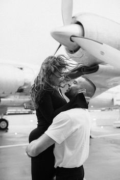 a man and woman standing in front of an airplane on the tarmac with their arms around each other