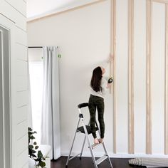 a woman standing on a ladder painting the wall with white paint in her living room