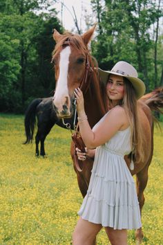 a woman in a white dress and cowboy hat standing next to a brown horse on a lush green field