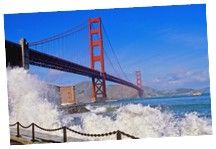 the golden gate bridge over looking the ocean with waves crashing against it and buildings in the background