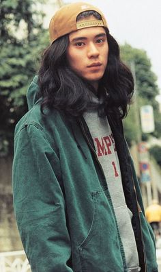 a man with long hair wearing a green jacket and brown hat standing in the street