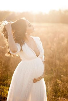 a pregnant woman in a white dress standing in a field with her hands on her head