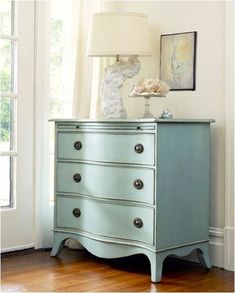 a blue dresser sitting in front of a window next to a lamp and painting on the wall