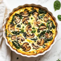 a quiche with spinach, cheese and mushrooms in a white dish on a marble surface