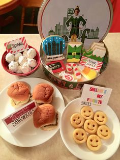 two plates with hamburgers and mini donuts on them next to a tin of cookies