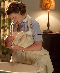 a woman holding a baby in her arms while standing next to a table with a lamp on it