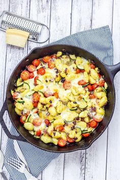 a skillet filled with pasta and vegetables on top of a wooden table next to cheese