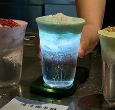 a person holding a glass with some food in it on a table next to two cups