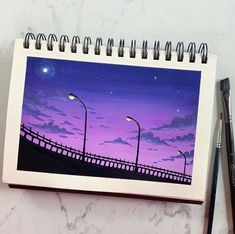 a notebook with a drawing of a purple sky and street lights on the cover, next to a pair of black pencils