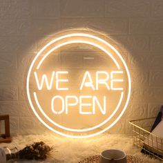 a neon sign that says we are open in front of a white brick wall with a basket on the floor