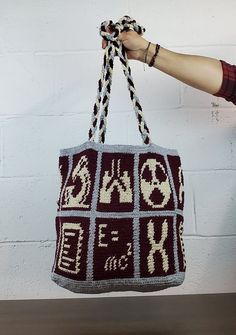 a person holding up a knitted bag with letters and numbers on it, in front of a white brick wall