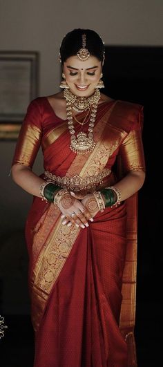 a woman in a red and gold saree smiles while holding her hands on her chest