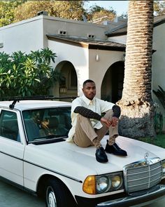 a man sitting on the hood of a white car in front of a palm tree