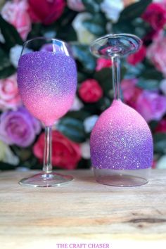 two wine glasses with pink and purple glitter on them sitting next to each other in front of flowers