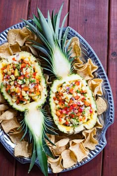 two pineapples with salsa and chips on a metal plate next to a wooden table