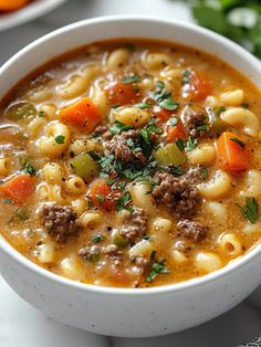 a white bowl filled with pasta and meat soup on top of a table next to carrots