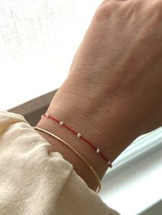 a woman's hand wearing a red string bracelet with white beads and a gold plated clasp