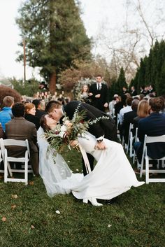 a bride and groom kissing in front of an outdoor wedding ceremony with guests seated on lawn chairs