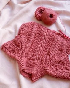 a pink knitted sweater and ball of yarn on a white bed sheet with two balls of thread