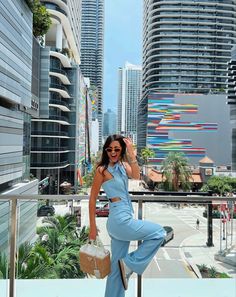 a woman in blue jumpsuits and sunglasses standing on a balcony with buildings behind her