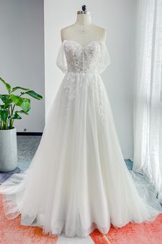 a white wedding dress on display in front of a large potted plant and window