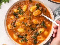 a person holding a spoon over a bowl of stew with dumplings and spinach