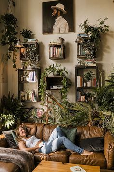 a woman laying on top of a brown couch in a living room filled with plants