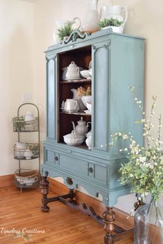 a blue china cabinet with dishes on top and flowers in vases next to it