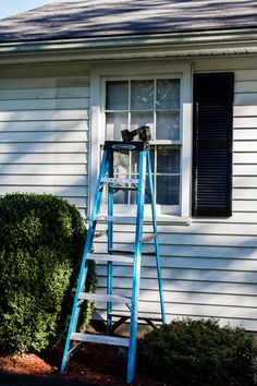 a blue ladder sitting in front of a white house next to a window with shutters