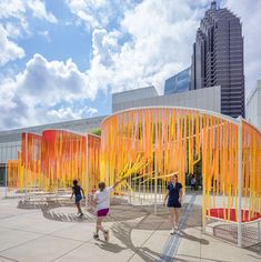 three people are standing in front of an art installation with orange sticks on it and buildings in the background