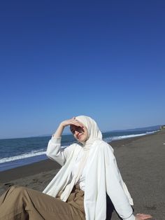 a woman sitting on the beach with her head in her hands and wearing a hijab