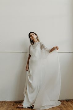 a woman standing in front of a wall wearing a white dress and holding her arms out