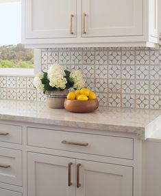 a bowl of lemons and some white flowers on a kitchen counter with tile backsplash