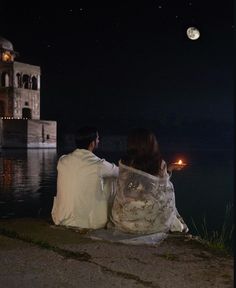 a man and woman sitting on the ground next to water at night