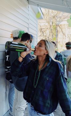 a woman drinking from a bottle while standing next to other people on the side of a house