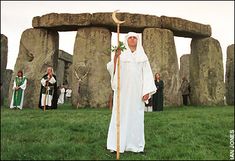 a man dressed in white holding a staff standing next to stonehenge with people around him