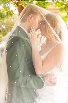 a bride and groom standing under a tree in their wedding day attire, kissing each other