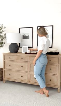 a woman standing on top of a dresser next to a lamp and framed pictures in a living room