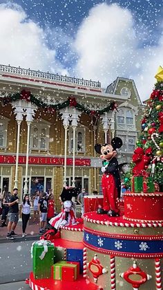 mickey mouse on top of a christmas tree in front of a building with people standing around it