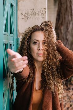 a woman with curly hair leaning against a blue door and pointing to the side while wearing a brown jacket