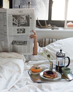 a person laying in bed reading a newspaper with breakfast on the table next to them