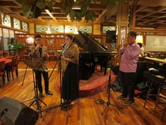 three people are standing in front of a piano and singing into microphones while another person stands behind them