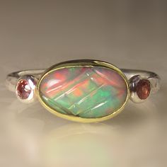 Beautiful, natural, solid carved, Ethiopian opal cabochon set in solid 18k gold bezel on hammered sterling silver band(approx. 2mm wide X 1.5mm thick - hammered from comfy oval wire). Fine silver backsheet underneath stone which has been blackened to bring out the fire of the opal. 2.5mm peachy pink faceted sapphires set in sterling silver on each side of opal. The opal weighs 0.91cts and measures 6.7 X 10.2mm. All metal is from recycled sources. This ring is a size 7 1/4 and I can stretch to a Luxury Handmade Opal Ring For Women, Luxury Heirloom Cabochon Opal Ring, Luxury Oval Cabochon Opal Ring For Formal Occasions, Boulder Opal Ring, Opal Band, Ethiopian Opal Ring, Ring Opal, Pink Sapphire Ring, Hammered Sterling Silver