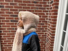 a young boy wearing a furry hat and scarf on his head while standing in front of a brick building