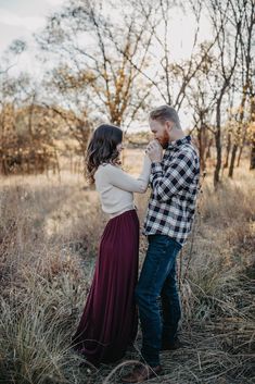 an engaged couple standing in the middle of a field with tall grass and trees behind them